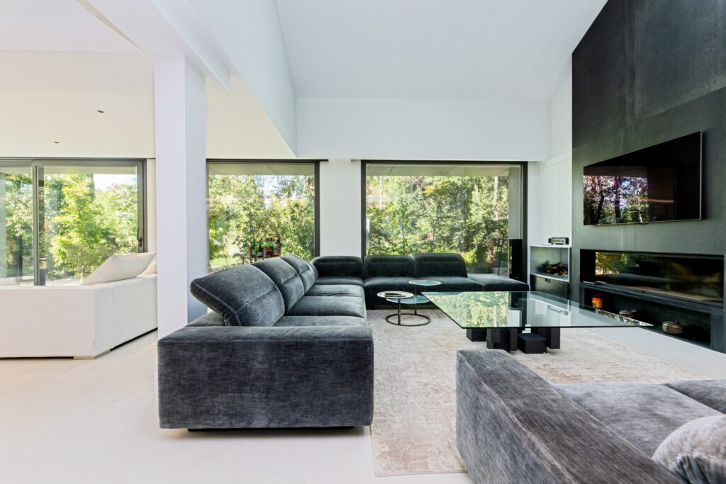 Salón contemporáneo con sofá en tonos grises, grandes ventanales al jardín y chimenea moderna integrada en pared oscura