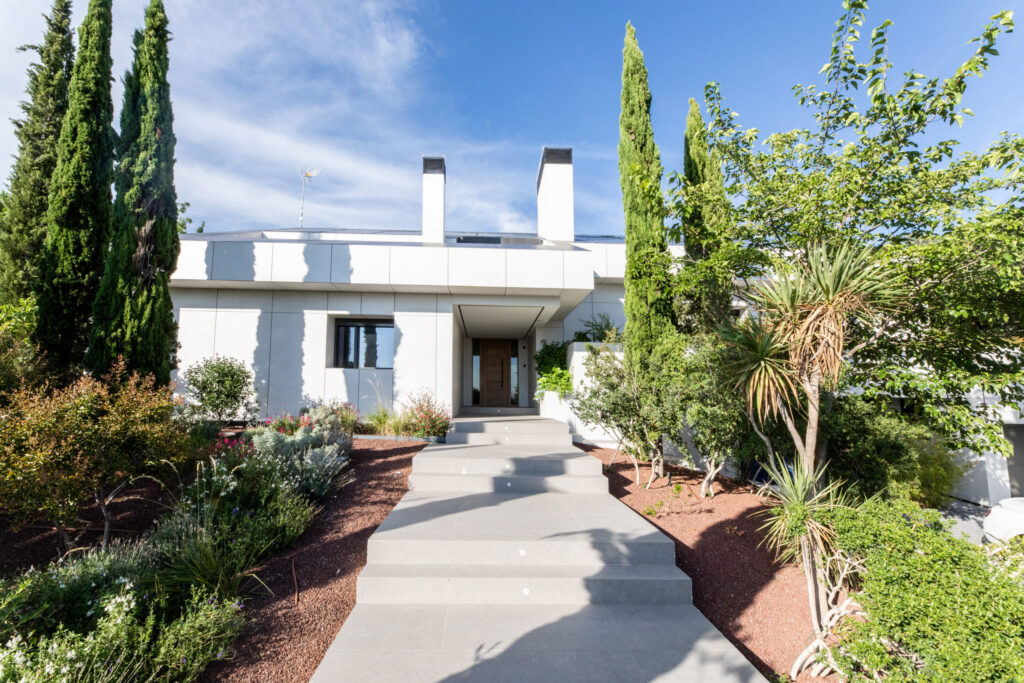 Entrada de vivienda moderna con fachada blanca, jardín paisajístico y acceso peatonal