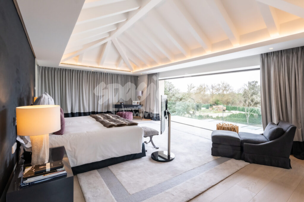 Dormitorio principal de vivienda de lujo con techo abuhardillado, iluminación indirecta y ventanales al jardín
